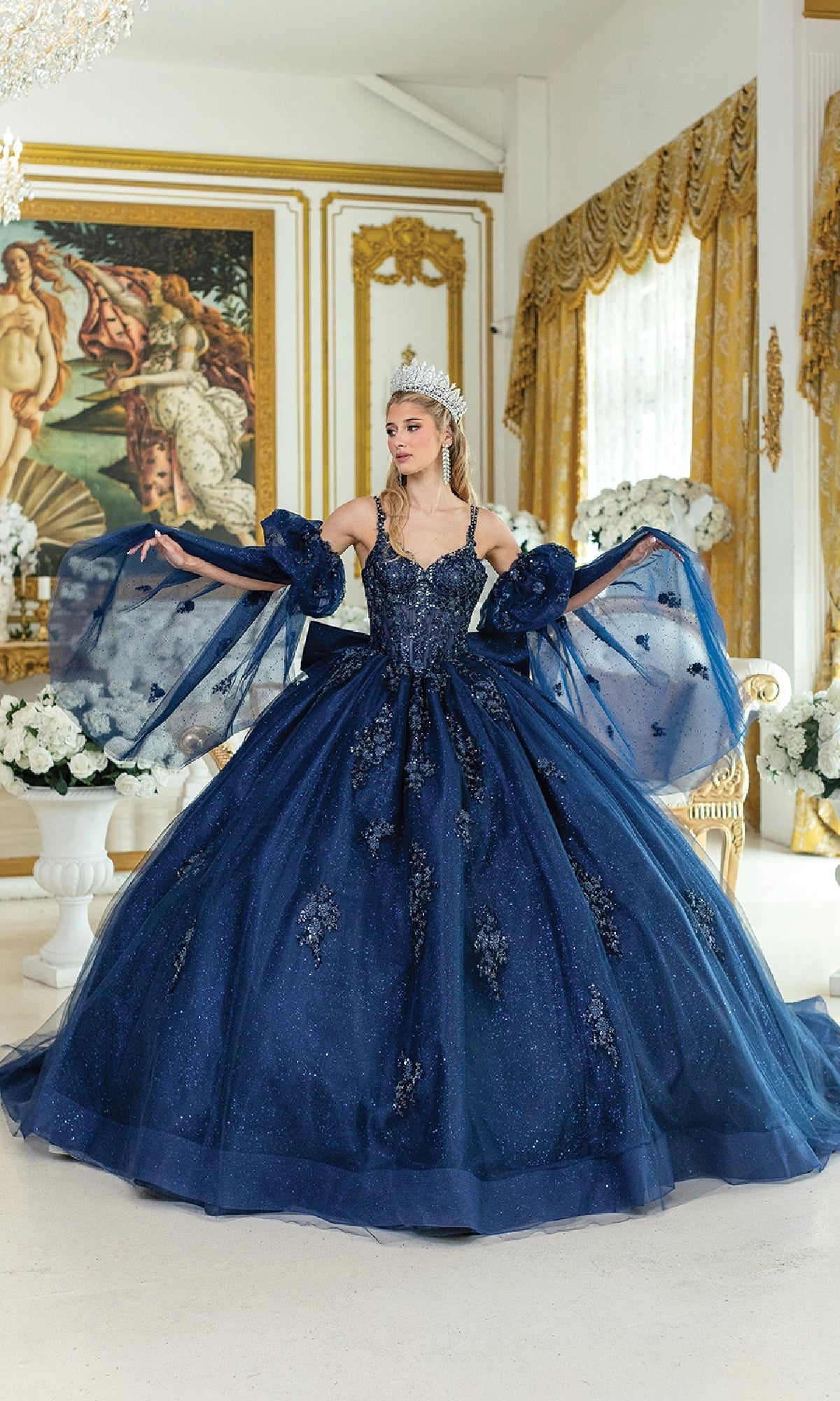 A woman in the Blue Quinceanera Dress 5037 By Dancing Queen and a tiara poses indoors, surrounded by white flowers and ornate gold-trimmed decor with classical paintings in the background.